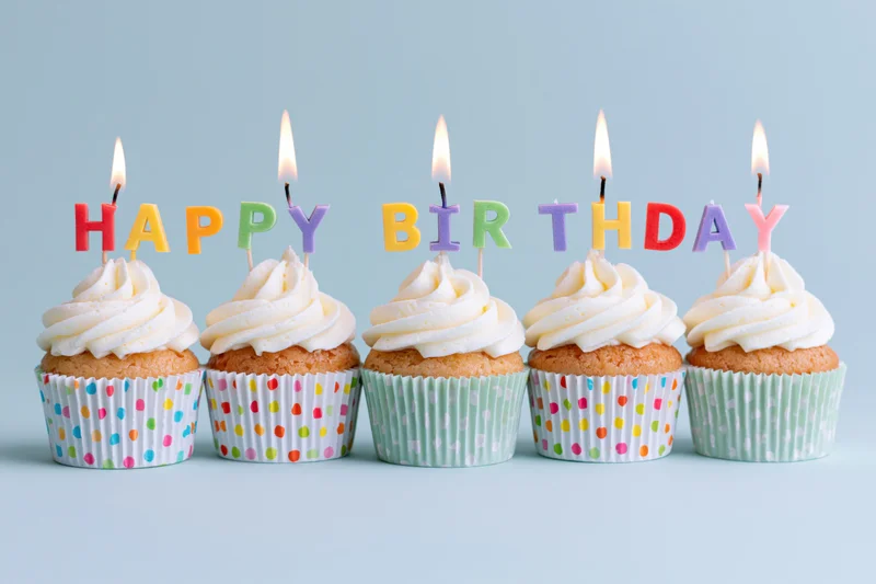 Five cupcakes with white frosting and lit letter candles spelling happy birthday in colorful wrappers on a light blue surface