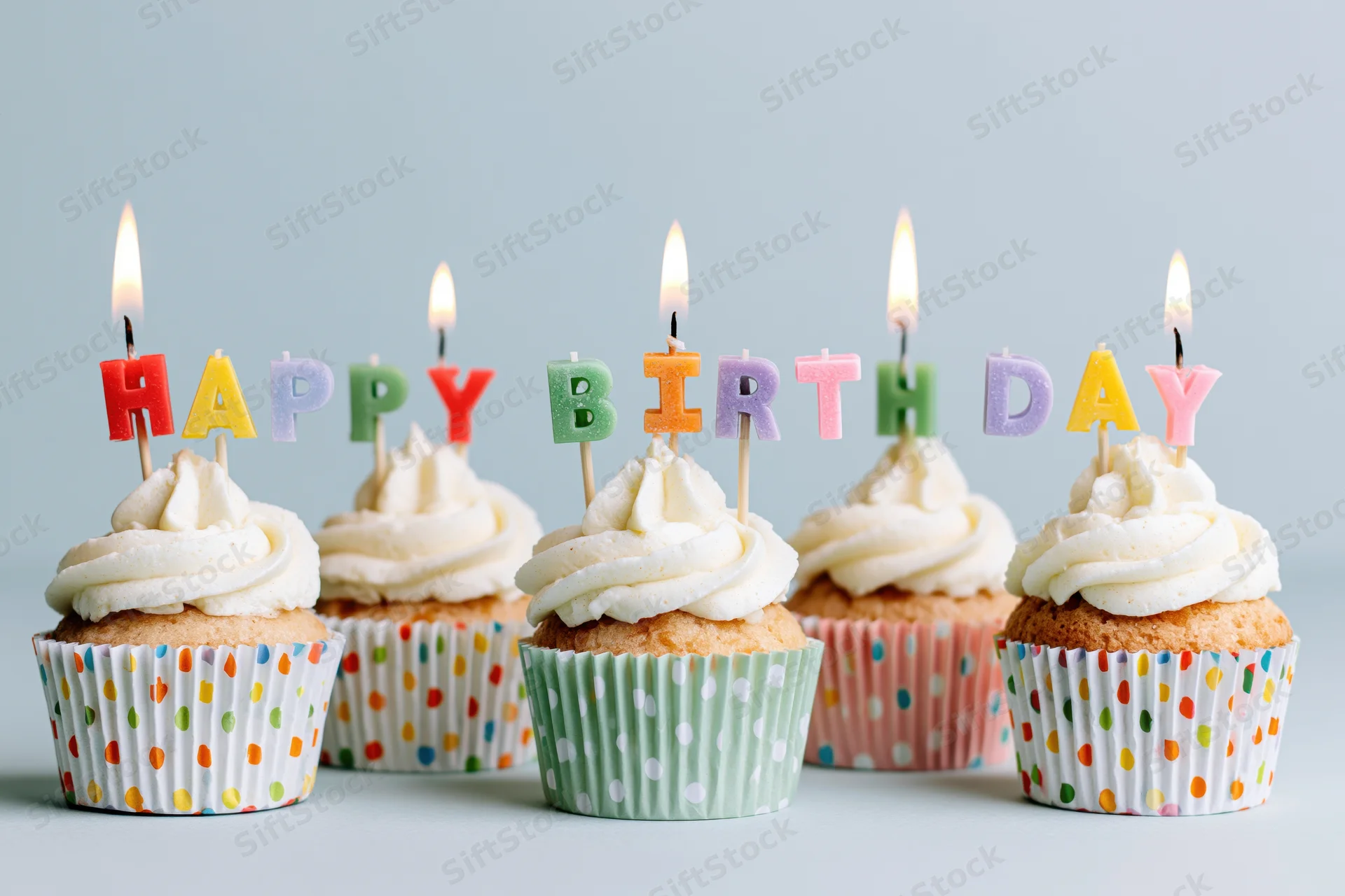 Five cupcakes with white frosting and lit letter candles spelling happy birthday in colorful paper liners on a light surface
