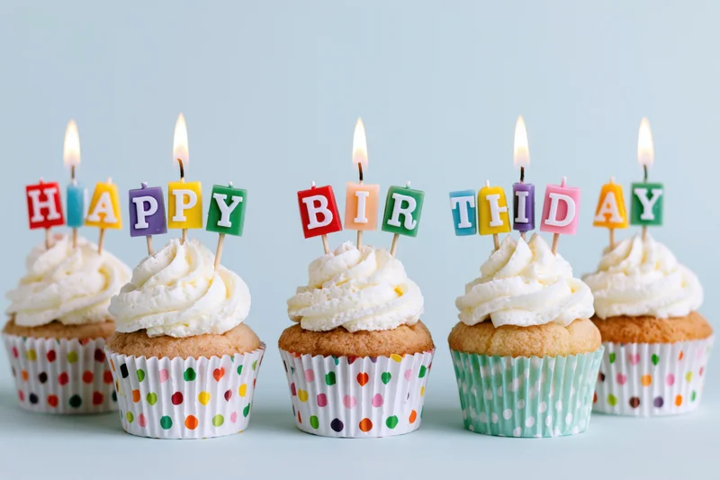 Five cupcakes with white frosting and lit letter candles spelling happy birthday in colorful paper liners on a light background