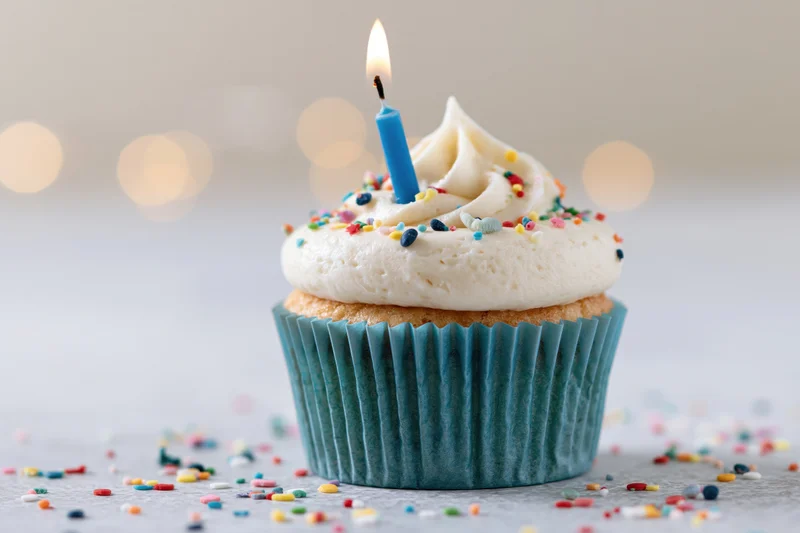 Single birthday cupcake with lit blue candle and sprinkles on white surface
