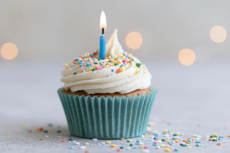 Single birthday cupcake with lit blue candle and colorful sprinkles on white surface