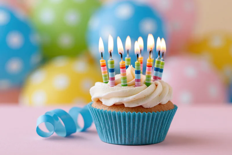 Birthday cupcake with lit candles on pink surface, colorful balloons in background