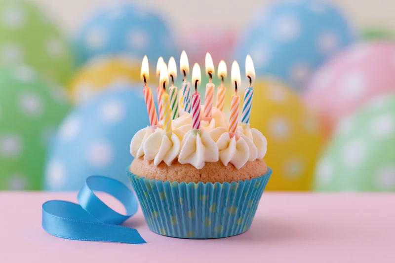 Birthday cupcake with lit candles on pink surface, blue ribbon beside, colorful balloons in background