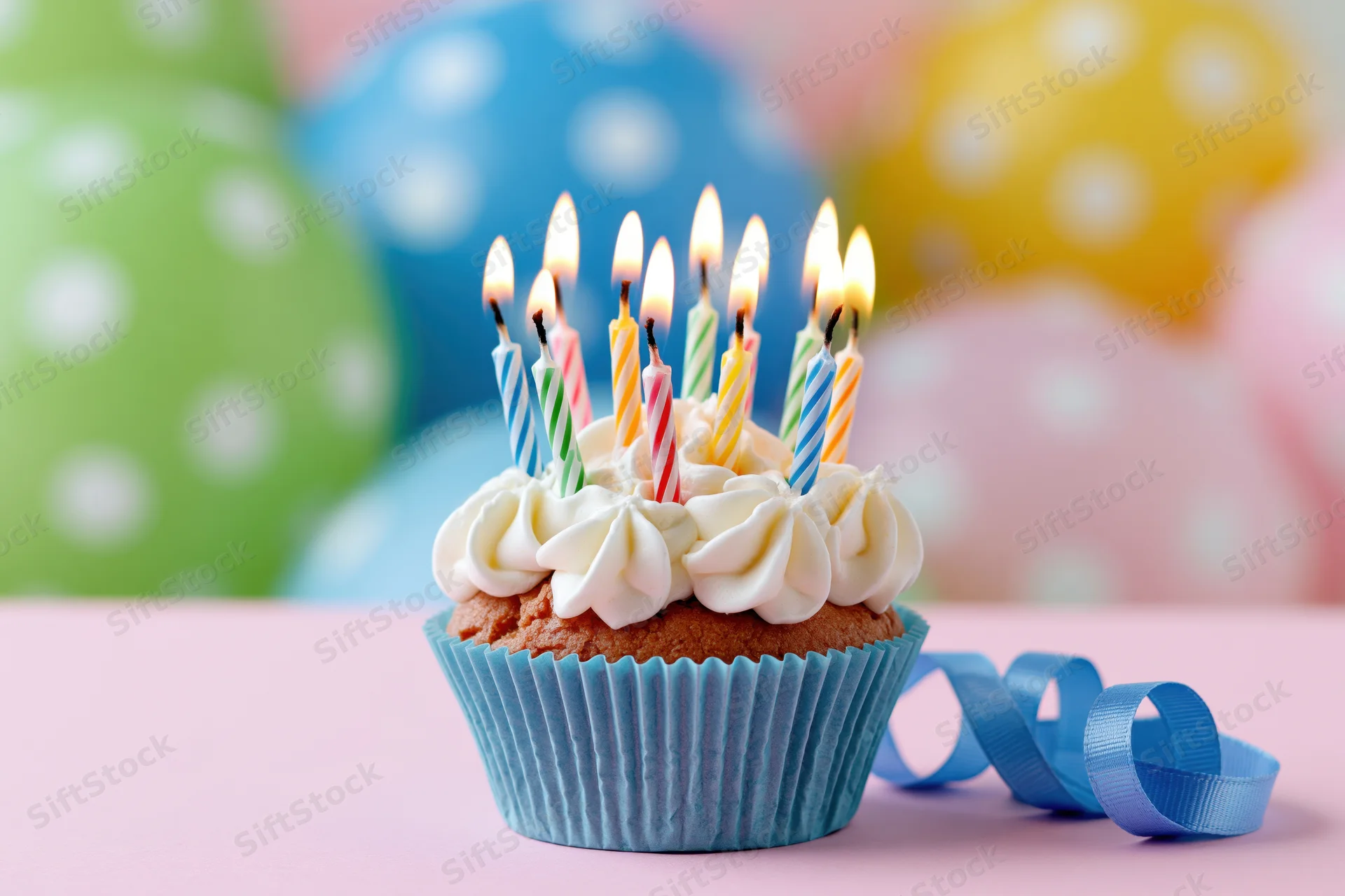 Birthday cupcake with lit candles and frosting on a pink surface, surrounded by colorful polka-dotted balloons and a blue ribbon