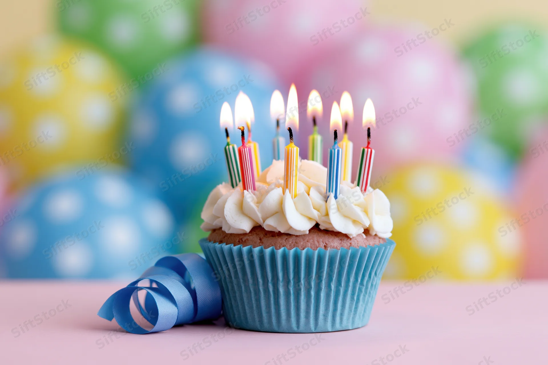 Birthday cupcake with lit candles and ribbon on a pastel surface, colorful balloons in the background
