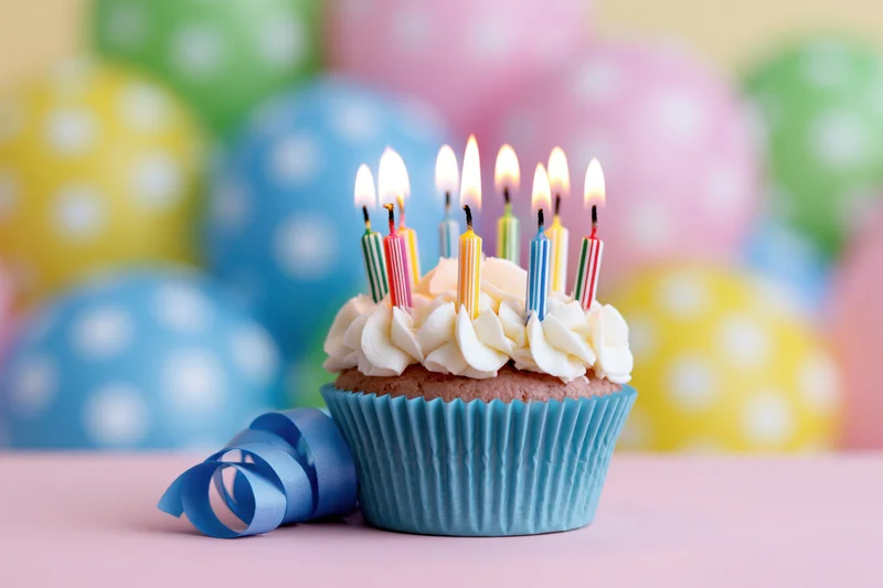 Birthday cupcake with lit candles and ribbon on a pastel surface, colorful balloons in the background