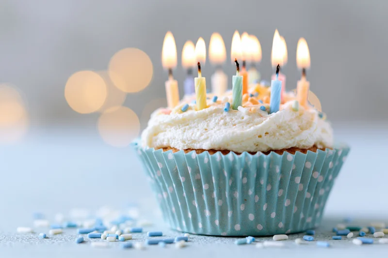 Single birthday cupcake with lit candles and sprinkles in a blue polka-dot paper liner on a light surface