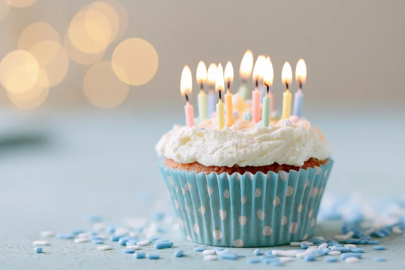 A birthday cupcake with lit candles and white frosting in a blue polka-dot paper liner on a light surface with scattered sprinkles and bokeh lights in the background