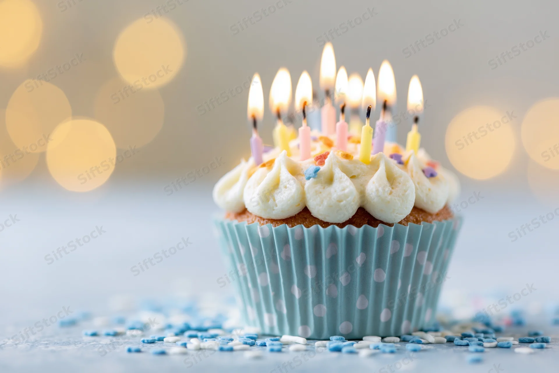 Birthday cupcake with lit candles in a blue polka-dot paper liner surrounded by sprinkles on a light surface with bokeh background