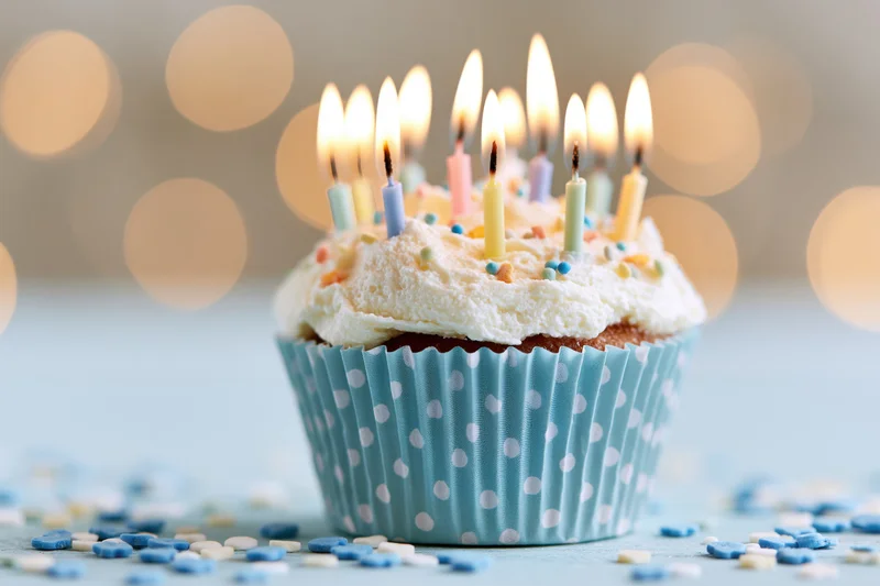 Single birthday cupcake with lit candles in a blue polka-dot paper liner on a surface with scattered sprinkles and bokeh lights behind