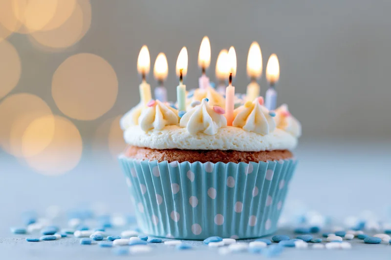 Birthday cupcake with lit candles in a blue polka-dot paper liner surrounded by sprinkles on a soft blurred background