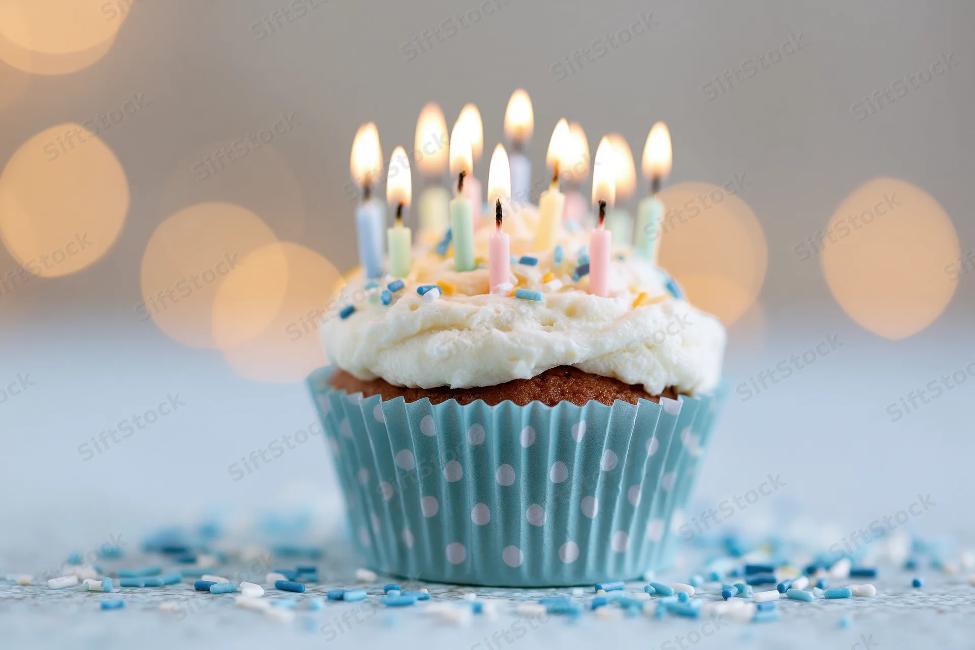 Single birthday cupcake with lit candles and sprinkles in a blue polka-dot paper liner on a light surface with bokeh lights behind