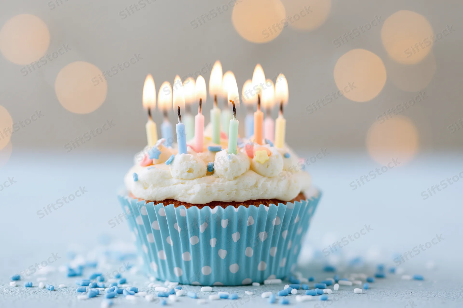 Single birthday cupcake with lit candles in blue polka-dot liner on a light surface with scattered sprinkles and bokeh background