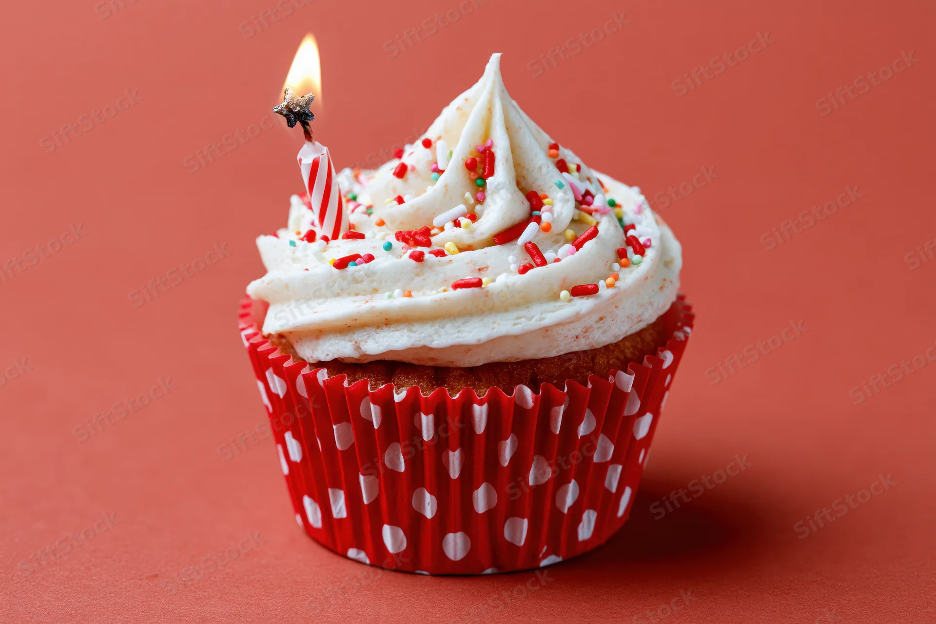 Single birthday cupcake with lit candle and sprinkles in red polka-dot paper liner on coral background