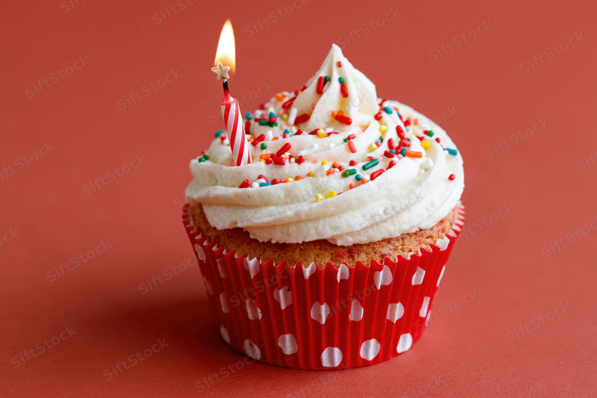Birthday cupcake with lit candle and sprinkles in red polka-dot paper liner on red background