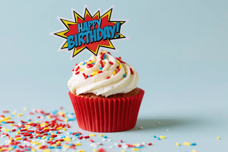 Single birthday cupcake with comic-style topper and sprinkles on light blue surface