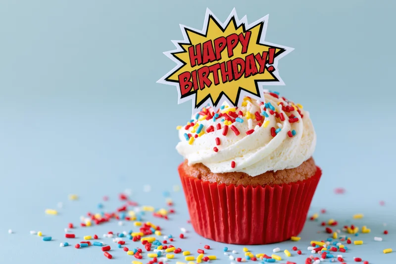 Single birthday cupcake with white frosting and colorful sprinkles in red liner, topped with comic-style happy birthday topper on light blue background