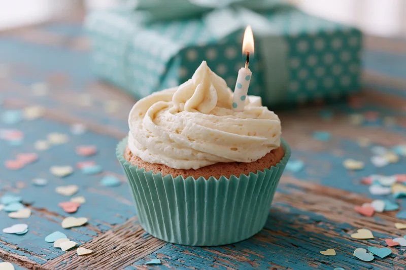 Birthday cupcake with lit candle and frosting swirl on wooden table surrounded by heart confetti and gift box