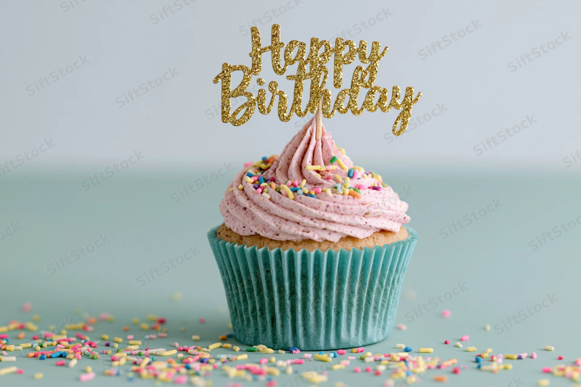 Single birthday cupcake with pink frosting and gold topper on teal surface surrounded by sprinkles