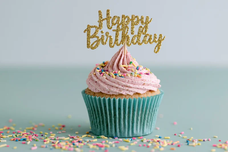 Single birthday cupcake with pink frosting and gold topper on teal surface surrounded by sprinkles
