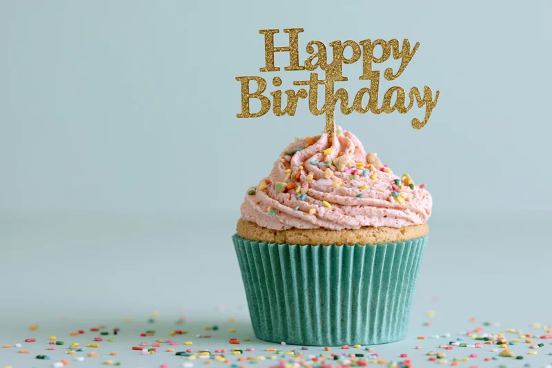 Single birthday cupcake with pink frosting and gold topper in teal wrapper on light blue surface with scattered sprinkles