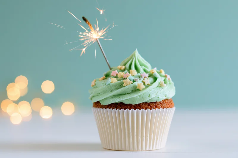 Single birthday cupcake with green frosting and sparkler on white surface against teal background with bokeh lights