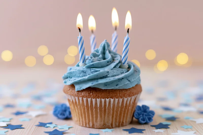 Single birthday cupcake with blue frosting swirl and lit candles on wooden surface with star confetti and bokeh lights