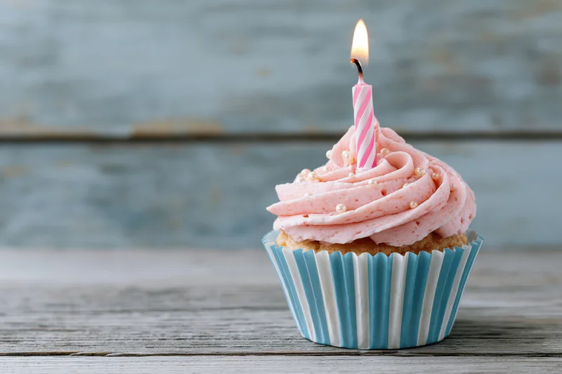 Single birthday cupcake with pink swirled frosting and lit candle on wooden table