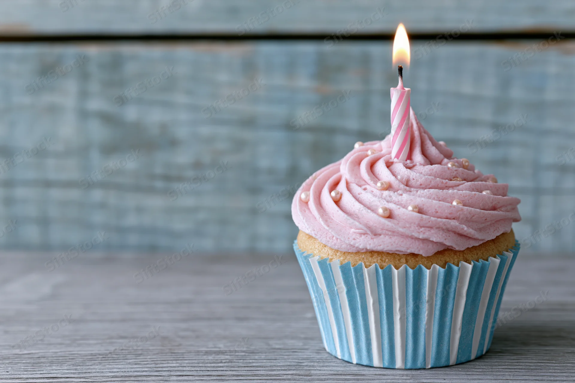 Single birthday cupcake with pink swirled frosting and lit candle on wooden table