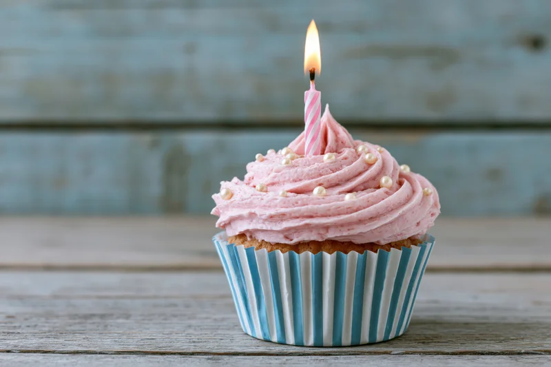 Single birthday cupcake with pink swirled frosting and lit candle on wooden table