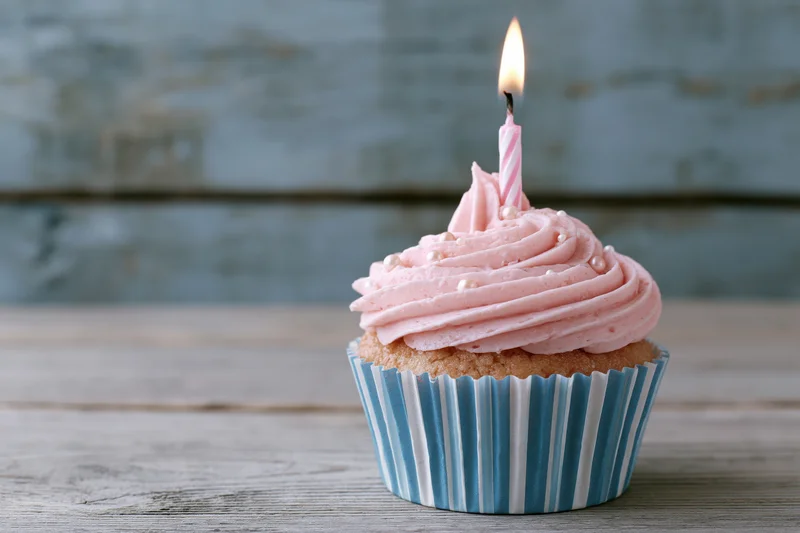 Single birthday cupcake with pink swirled frosting and lit candle on wooden table