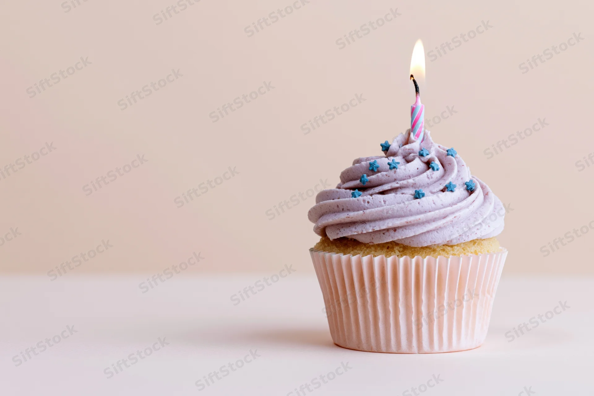 Single birthday cupcake with swirled lavender frosting and lit candle on a plain surface