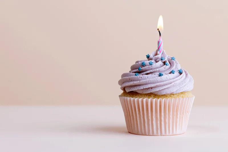 Single birthday cupcake with swirled lavender frosting and lit candle on a plain surface