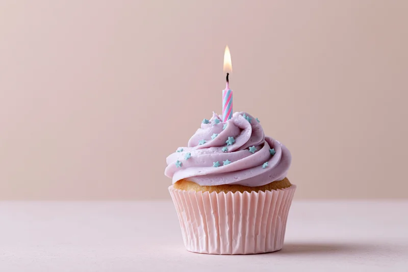 Single birthday cupcake with lit candle and lavender frosting on pink surface