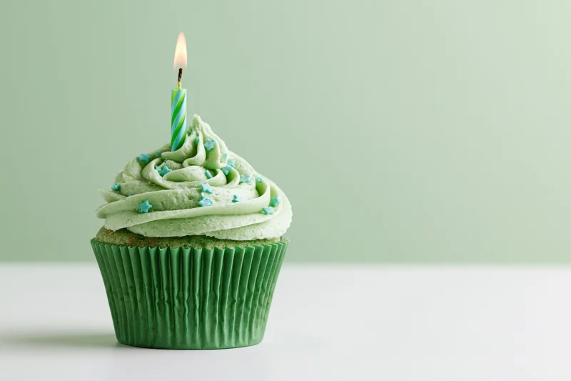 Single green-frosted cupcake with lit candle on white surface against pastel green background
