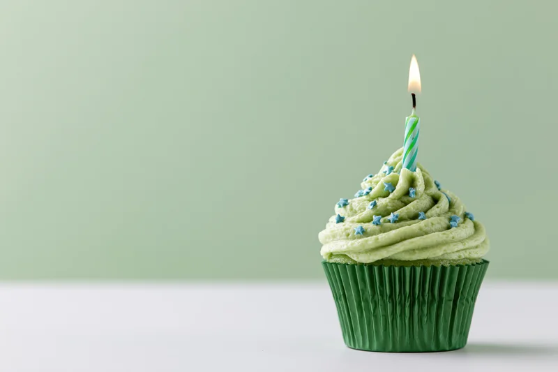 Single birthday cupcake with swirled green frosting and lit candle on white surface against green background