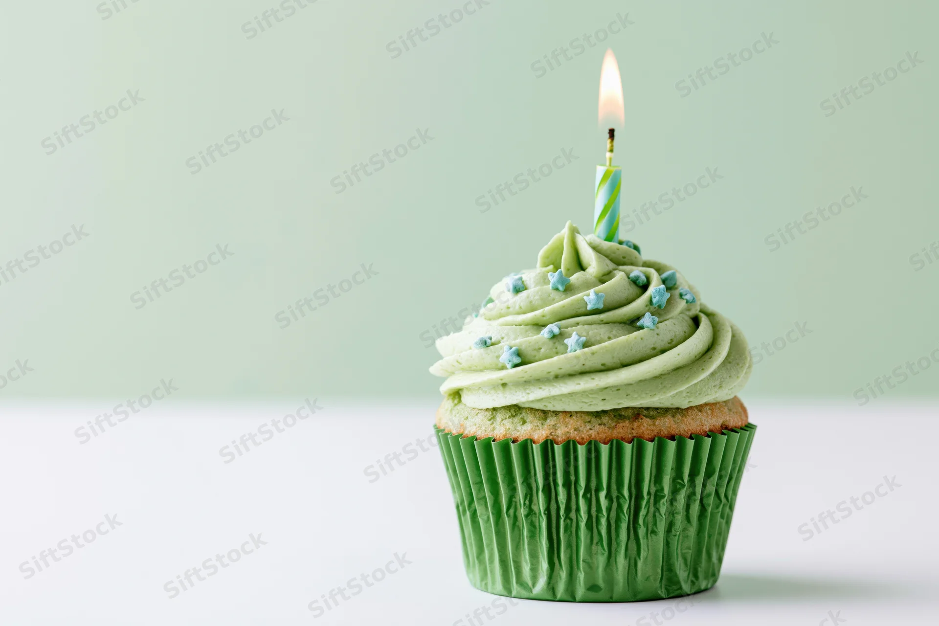 Single green-frosted cupcake with lit candle and star sprinkles on white surface