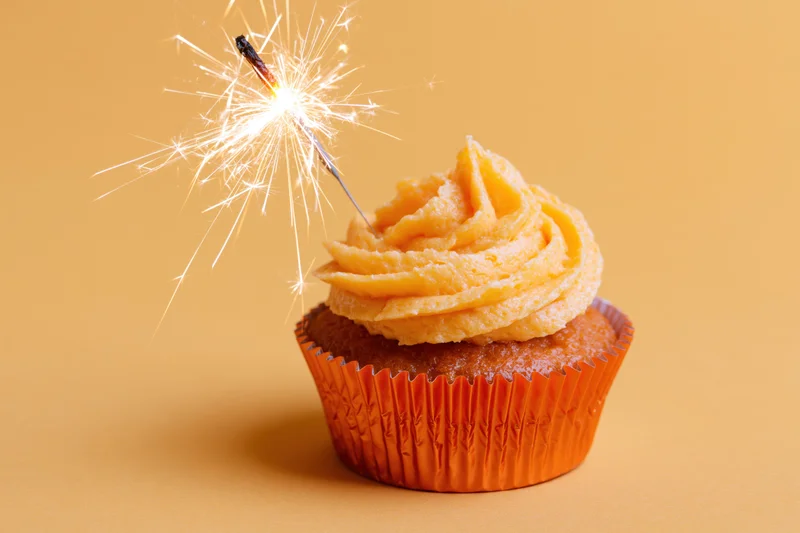 Single birthday cupcake with swirled orange frosting and lit sparkler on plain orange background