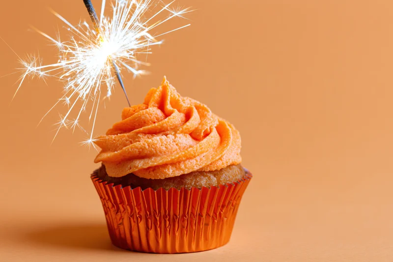 Orange-frosted cupcake with lit sparkler on warm background