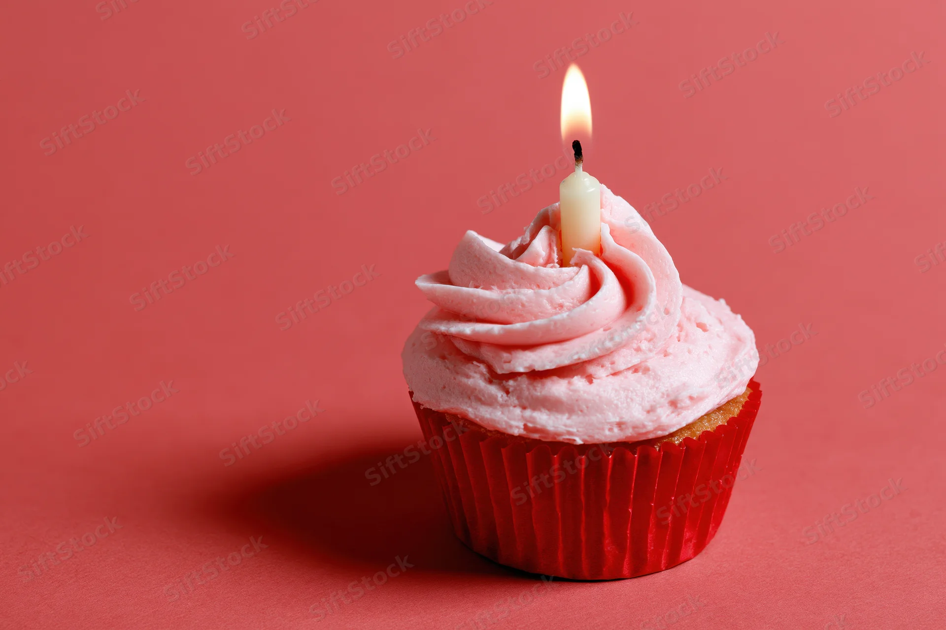 Single birthday cupcake with pink frosting and lit candle on red background