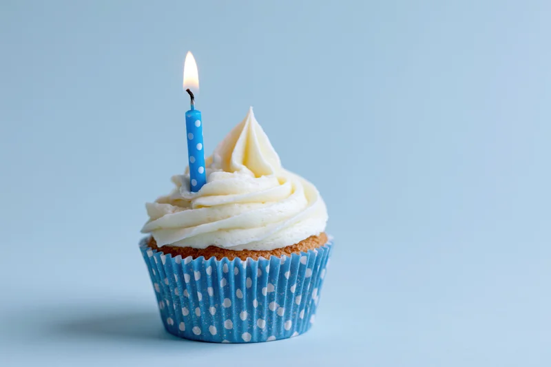 Single birthday cupcake with white frosting and one lit blue candle in a polka dot liner on a light blue background