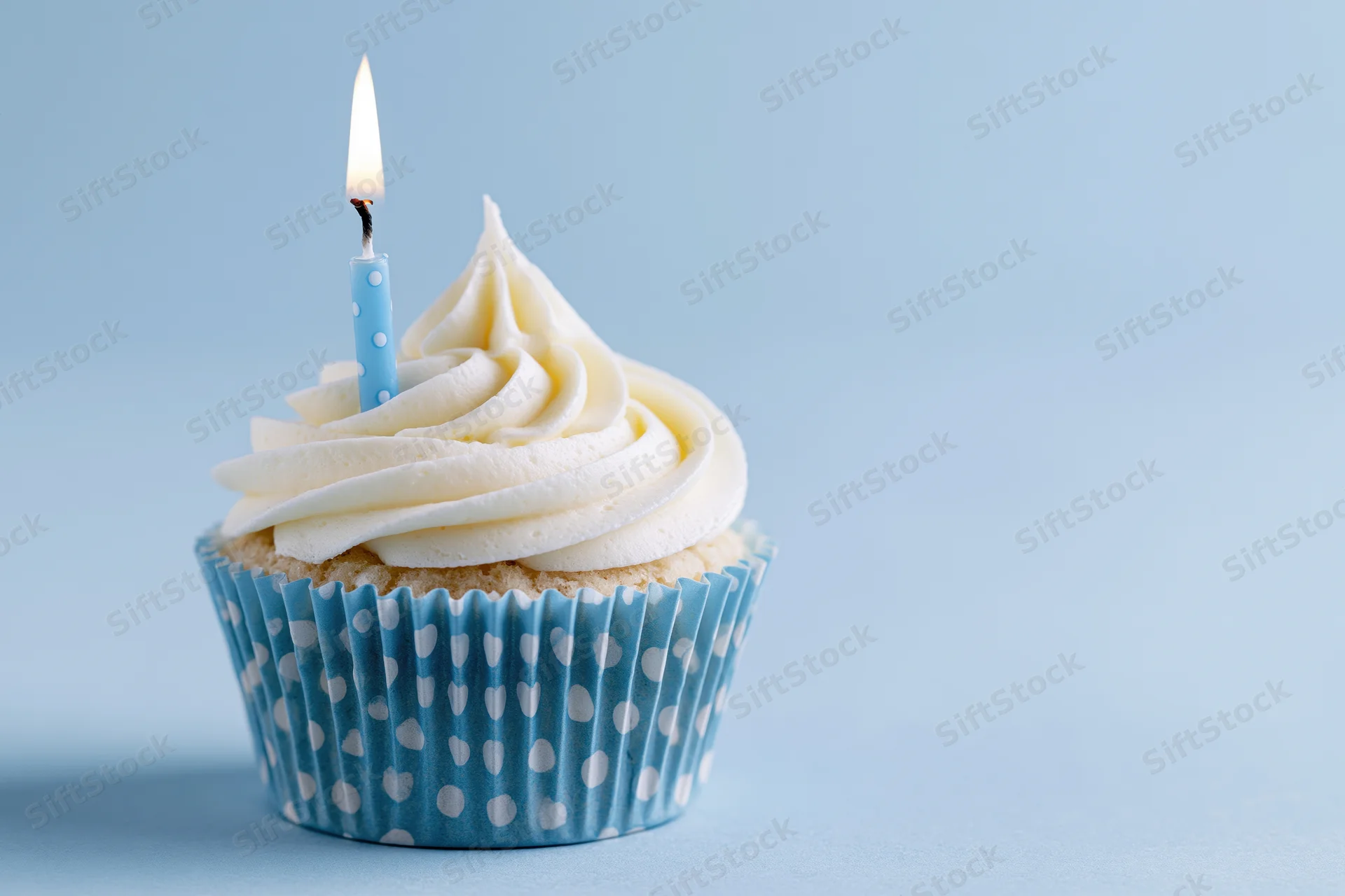 Single birthday cupcake with white frosting and one lit blue candle in a polka dot paper liner on a light blue background