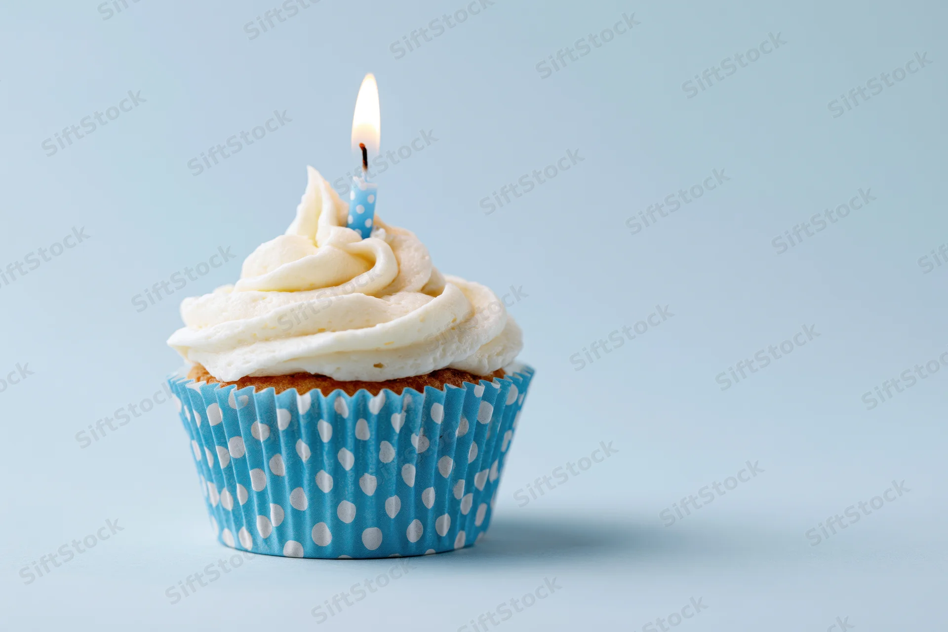 Single birthday cupcake with white frosting and lit candle in blue polka dot liner on light blue background