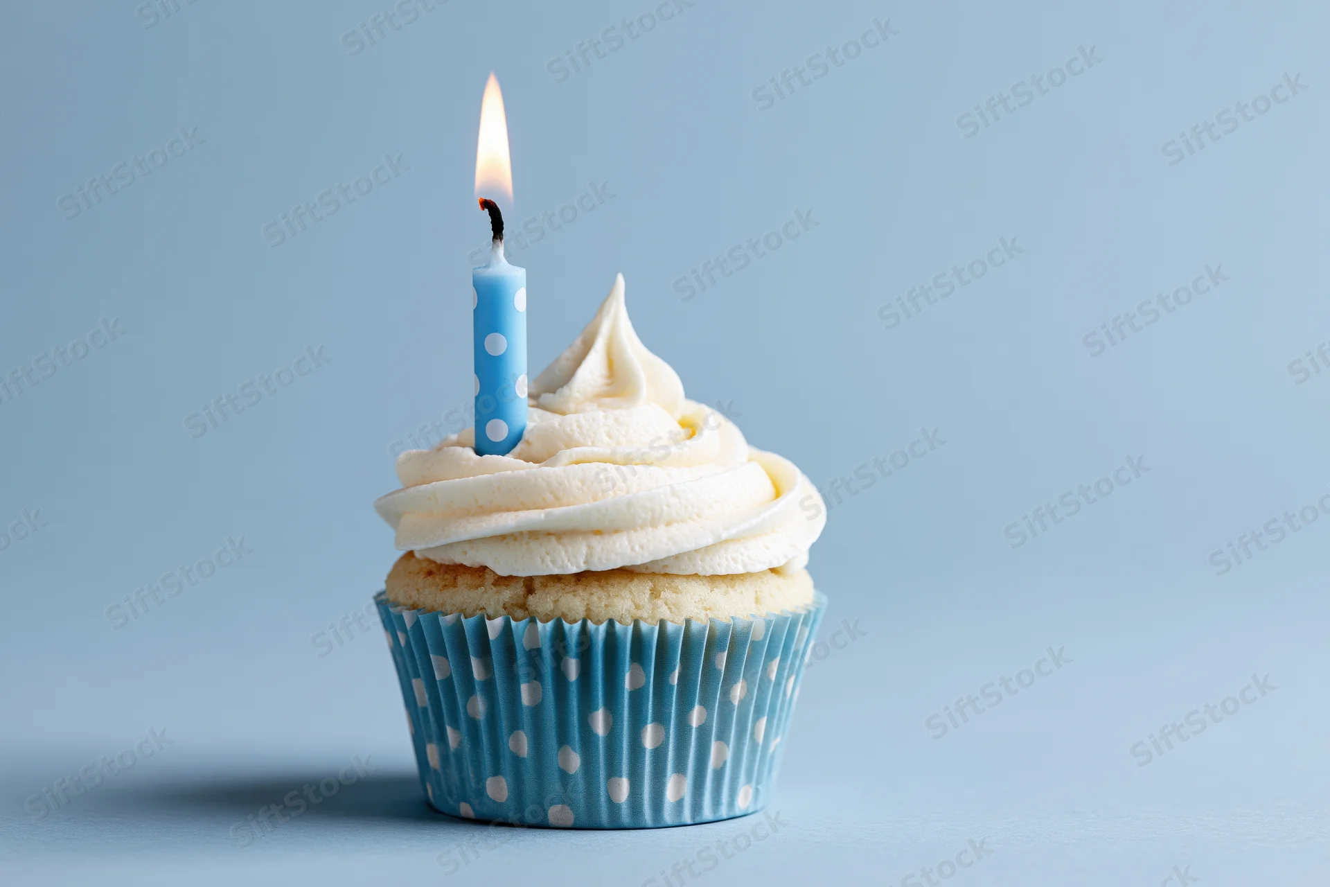 Single birthday cupcake with white frosting and one lit blue candle in a polka dot paper liner on a blue background