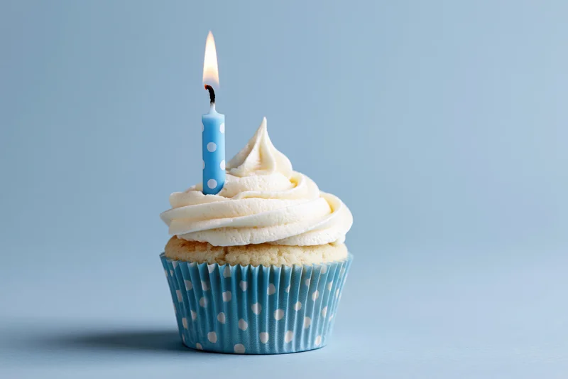 Single birthday cupcake with white frosting and one lit blue candle in a polka dot paper liner on a blue background