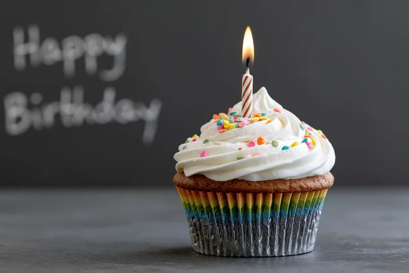 Single birthday cupcake with white frosting and lit candle on gray surface, rainbow liner, sprinkles, happy birthday text in background