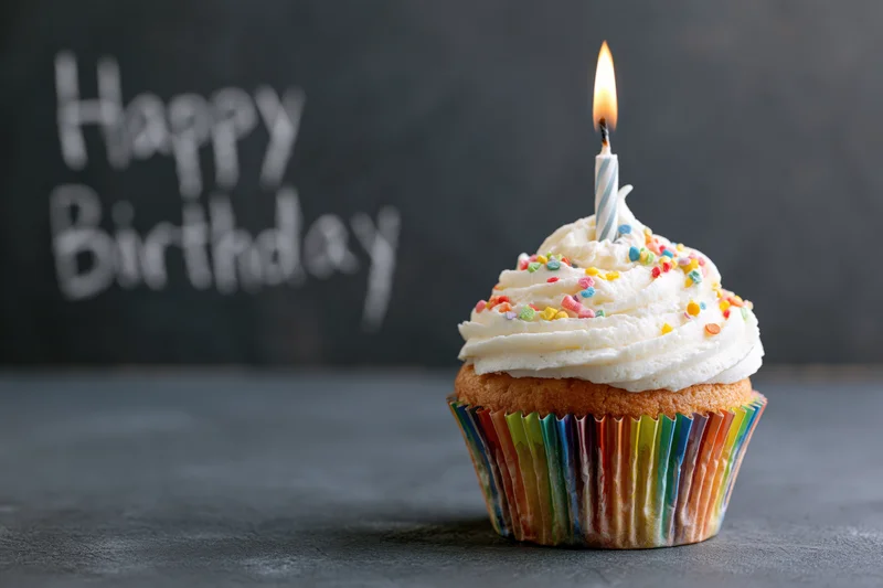 Single birthday cupcake with white frosting and lit candle on dark surface, rainbow liner, sprinkles, happy birthday message in background
