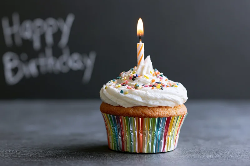 Single birthday cupcake with white frosting and lit candle on dark surface, colorful sprinkles and striped liner, "Happy Birthday" text in background