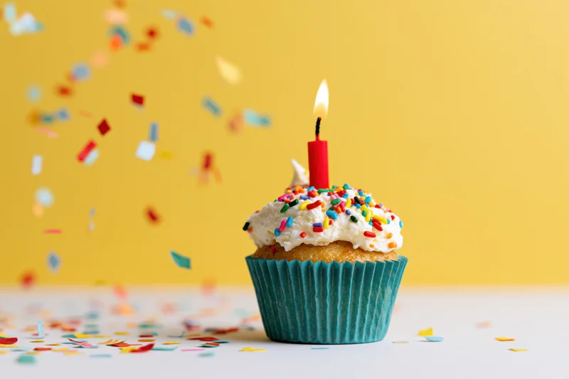 Birthday cupcake with lit candle and sprinkles on yellow background, confetti falling around it