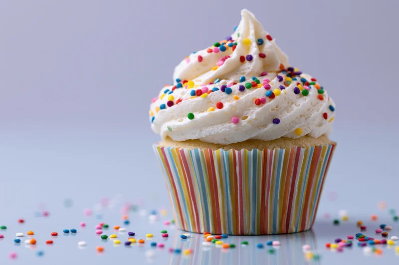 Cupcake with white frosting and colorful sprinkles in a rainbow-striped paper liner on a reflective surface